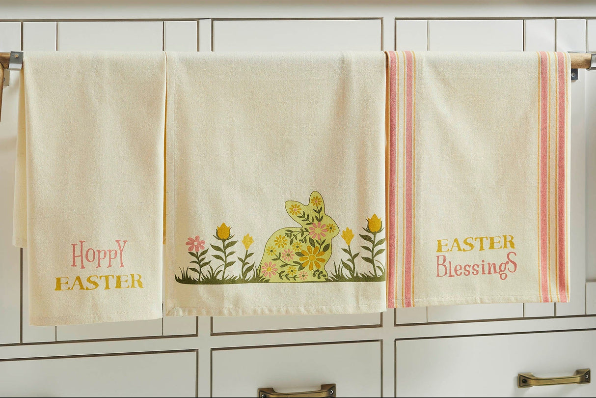 A set of three Easter-themed tea towels hanging on a rod, featuring floral bunny designs and Easter greetings in yellow, green, and pink colors on a cream background.