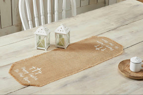 A light tan jute burlap table runner with the text 'Happy Easter He Has Risen' and simple crosses stenciled on it, placed on a wooden table with a white chair and decorative lanterns.