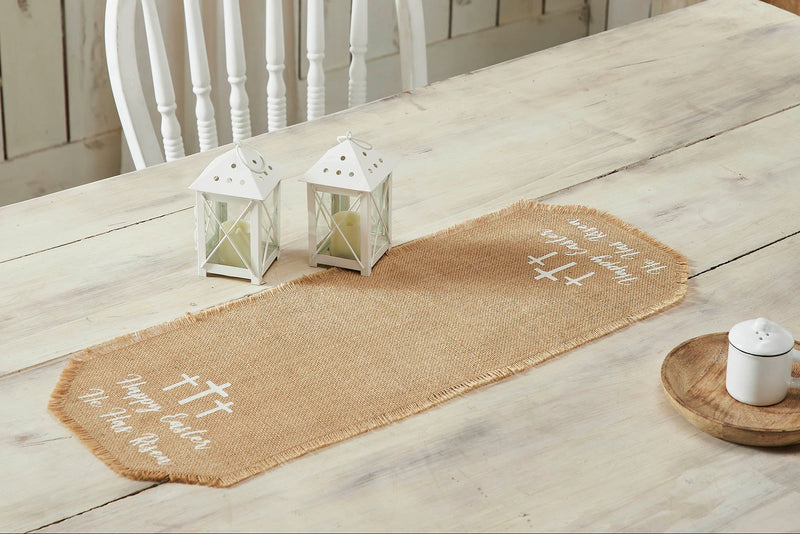 A light tan jute burlap table runner with the text 'Happy Easter He Has Risen' and simple crosses stenciled on it, placed on a wooden table with a white chair and decorative lanterns.